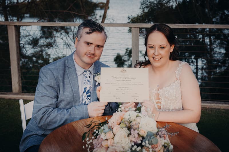 Rebecca and Dale sit at a wooden table outdoors at Sandstone Point Hotel — Rustic Arbour, holding and displaying their Certificate of Marriage with a floral centerpiece in front of them.