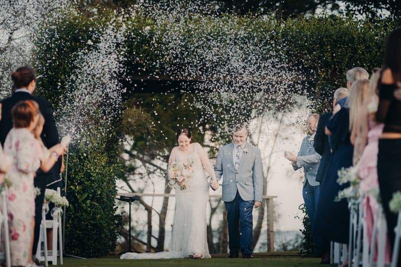 Bride Rebecca and groom Dale walk down the aisle hand in hand at Sandstone Point Hotel — Rustic Arbour as guests on both sides throw confetti.