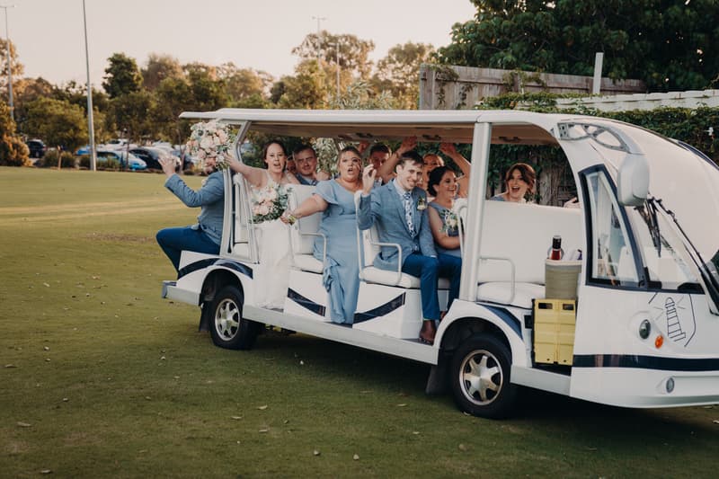 Rebecca the bride, Dale the groom, and their wedding party ride together in a white open-sided golf cart on the grass at Sandstone Point Hotel.