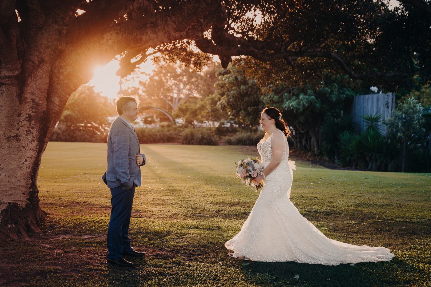 Rebecca in her wedding gown holding a bouquet walks toward Dale, who is standing near a large tree, at Sandstone Point Hotel during golden hour.