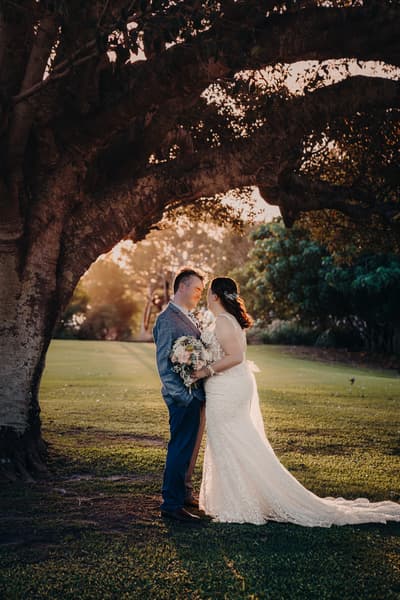 Rebecca the bride and Dale the groom stand facing each other under a large tree at Sandstone Point Hotel during their couple portraits session, with Rebecca holding a bouquet and the sun setting behind them.