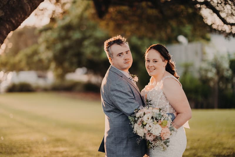 Rebecca and Dale pose together outdoors at Sandstone Point Hotel during their couple portraits session, with Rebecca holding a bouquet of flowers and Dale wearing a grey checked suit.