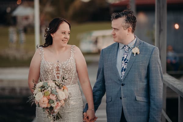 Rebecca the bride and Dale the groom hold hands and look at each other while standing outdoors at Sandstone Point Hotel. Rebecca wears a detailed lace wedding dress and holds a bouquet of flowers. Dale wears a blue checked suit with a floral tie and boutonniere.