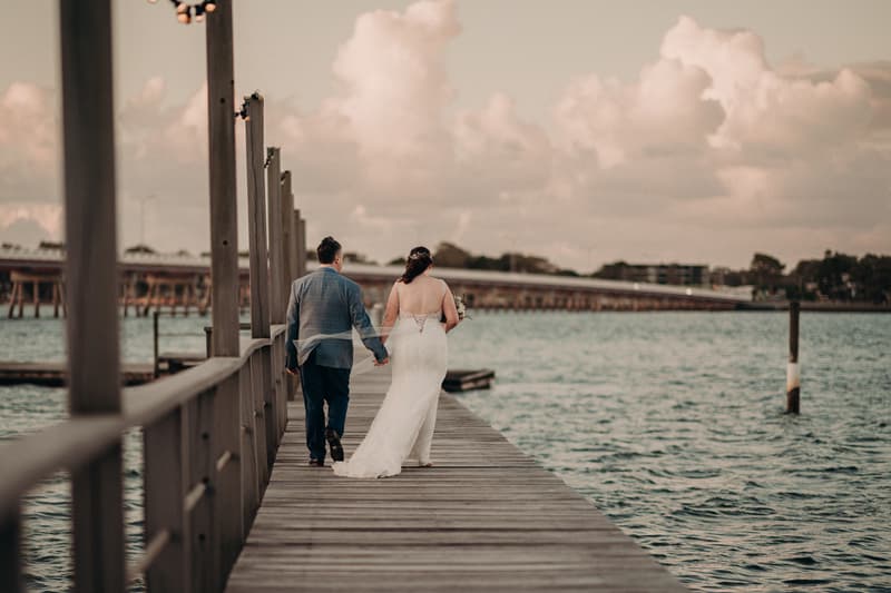 Bride Rebecca and groom Dale walk hand in hand along a wooden pier at Sandstone Point Hotel with water and a distant bridge in the background.