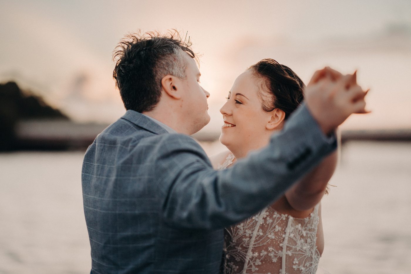 Rebecca and Dale pose for a couple portrait, holding hands and facing each other near the water at sunset. Wedding photographer and videographer by She Said Yes.