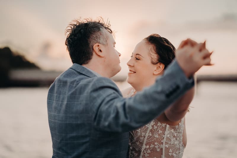 Rebecca and Dale pose for a couple portrait at Sandstone Point Hotel, holding hands and facing each other near the water at sunset.