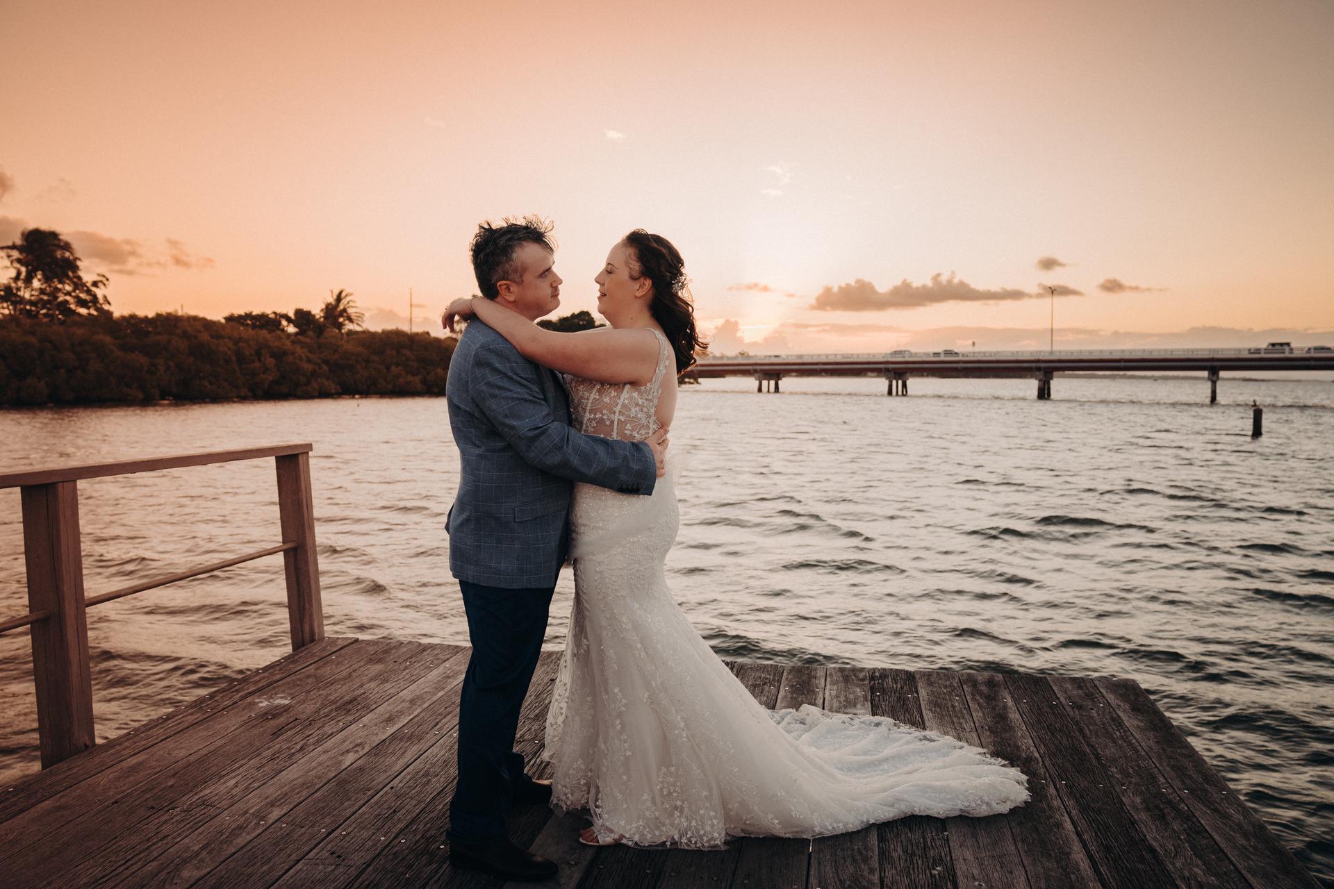 Rebecca and Dale embrace on a wooden jetty at Sandstone Point Hotel during sunset, with water and a distant bridge in the background.