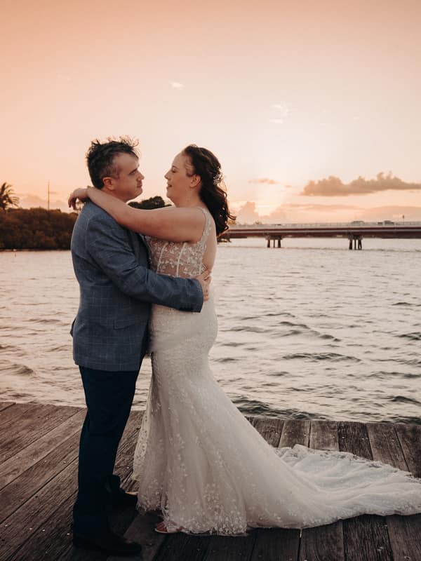 Rebecca and Dale embrace on a wooden jetty at Sandstone Point Hotel during sunset, with water and a distant bridge in the background.