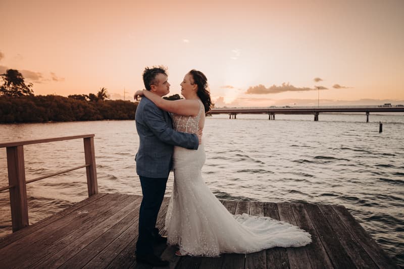 Rebecca and Dale embrace on a wooden jetty at Sandstone Point Hotel during sunset, with water and a distant bridge in the background.