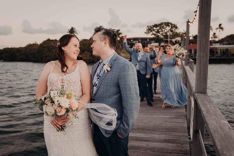 Rebecca the bride and Dale the groom stand close together on a wooden pier at Sandstone Point Hotel, with bridesmaids and groomsmen in blue attire lined up behind them.