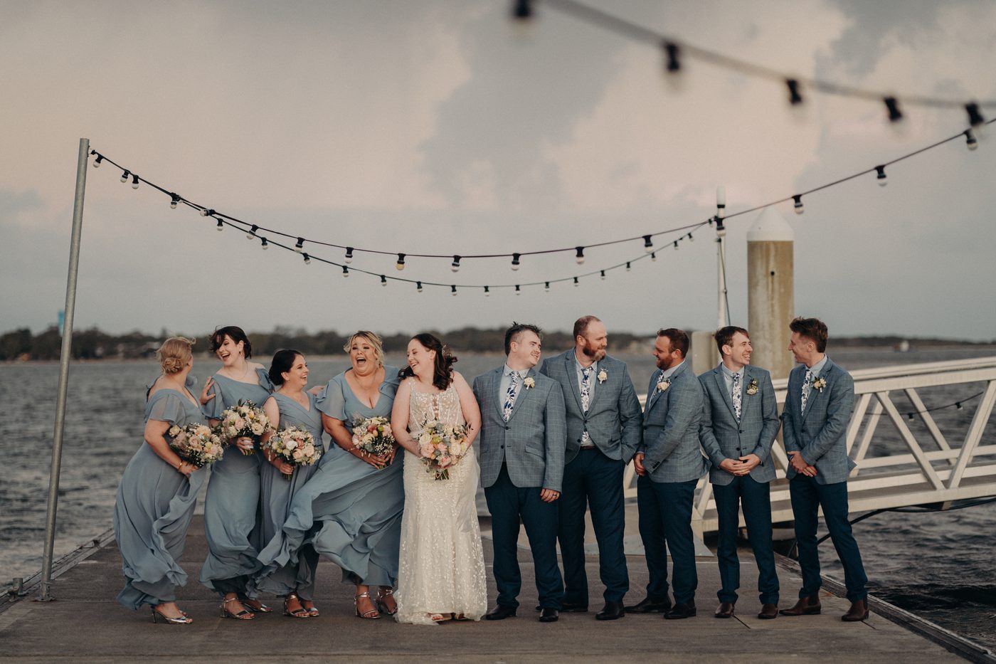 Rebecca the bride in a white gown stands with Dale the groom and their bridal party on a pier, with bridesmaids in blue dresses holding bouquets and groomsmen in blue suits with floral ties, under string lights by the water. Candid wedding photographer by She Said Yes.