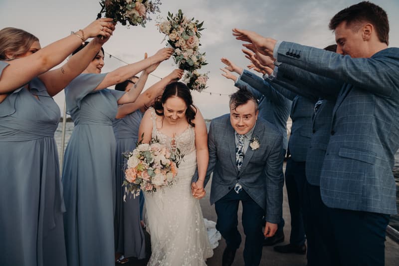 Rebecca the bride and Dale the groom crouch and hold hands while walking under an arch formed by bridesmaids in blue dresses holding bouquets and groomsmen in blue suits raising their arms at Sandstone Point Hotel.