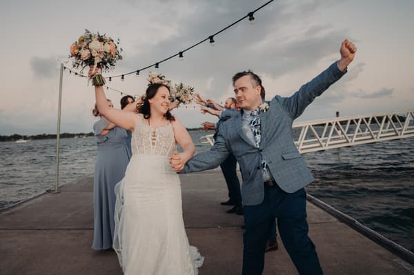 Rebecca the bride and Dale the groom hold hands and celebrate on a pier, with bridesmaids and groomsmen in the background holding bouquets and raising their arms. Wedding industry awards finalist by She Said Yes.
