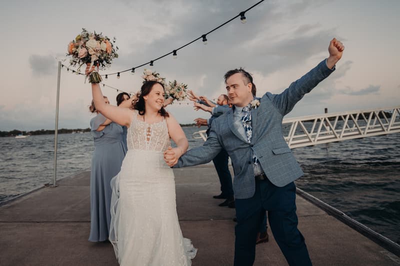 Rebecca the bride and Dale the groom hold hands and celebrate on a pier at Sandstone Point Hotel, with bridesmaids and groomsmen in the background holding bouquets and raising their arms.