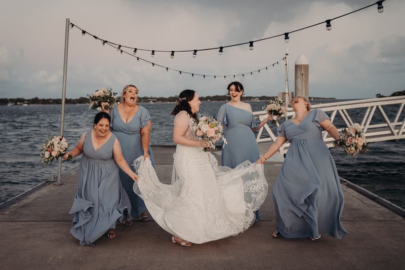 The bride Rebecca stands on a pier at Sandstone Point Hotel with four bridesmaids in blue dresses holding bouquets, all smiling and interacting with each other.