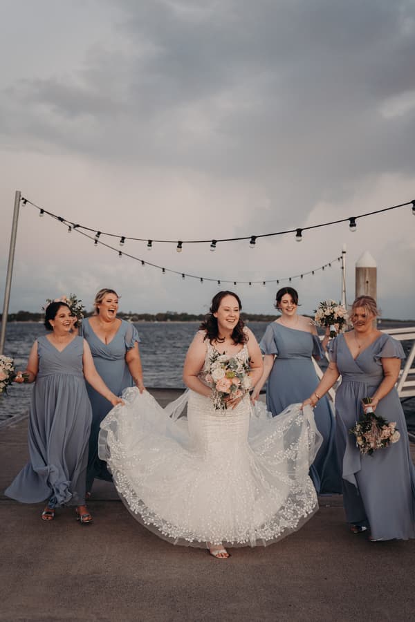 The bride Rebecca stands on a pier at Sandstone Point Hotel holding her bouquet, surrounded by four bridesmaids in blue dresses who are helping to hold her wedding gown train.