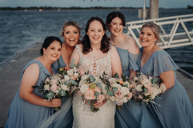 The bride Rebecca stands with four bridesmaids on a pier at Sandstone Point Hotel, each holding bouquets of flowers with the water visible in the background.