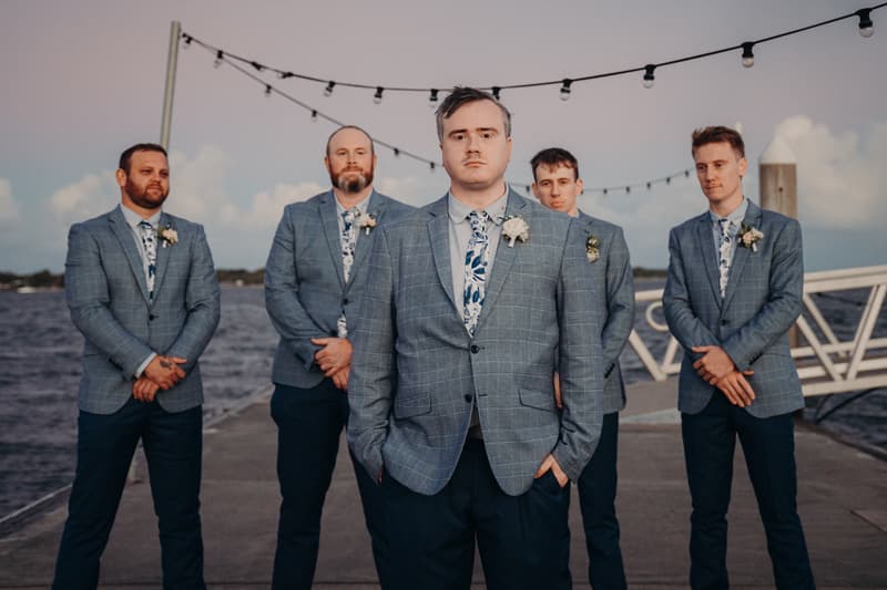 Five groomsmen stand on a pier at Sandstone Point Hotel, dressed in matching blue checkered suits with floral ties and boutonnieres, with string lights overhead and water in the background.