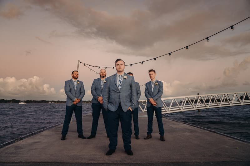 Dale and four groomsmen stand on a pier at Sandstone Point Hotel with water and a cloudy sky in the background, wearing matching blue suits with floral ties and boutonnieres.