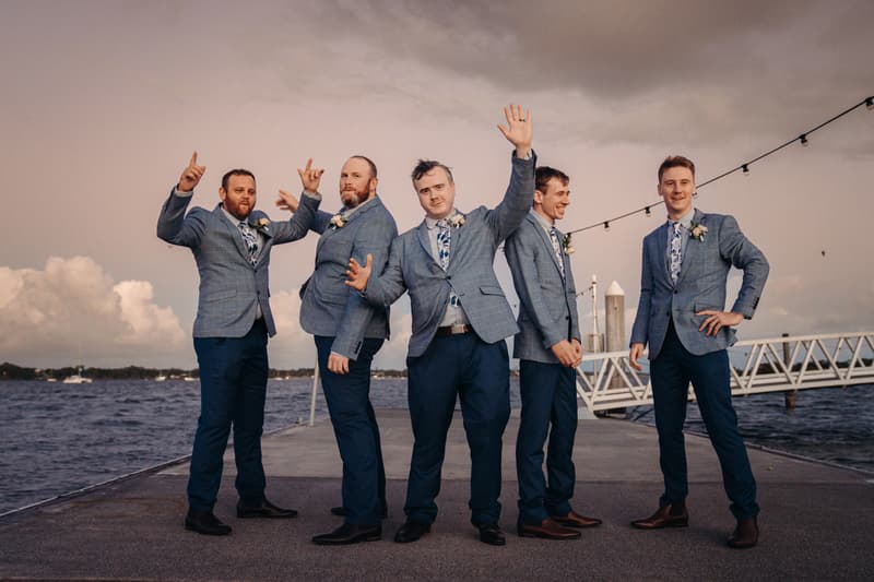Five groomsmen pose on a pier at Sandstone Point Hotel with water and a cloudy sky in the background.