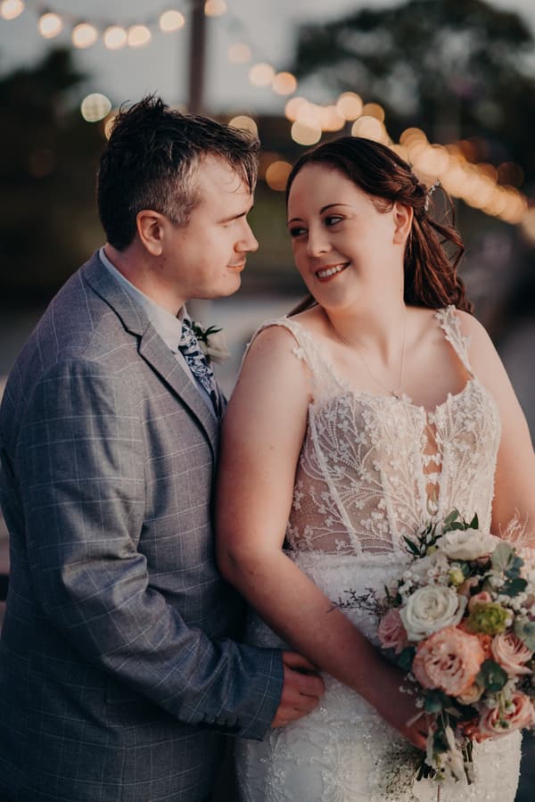 Rebecca and Dale pose for couple portraits at Sandstone Point Hotel, with Rebecca holding a bouquet of flowers and Dale standing close behind her.