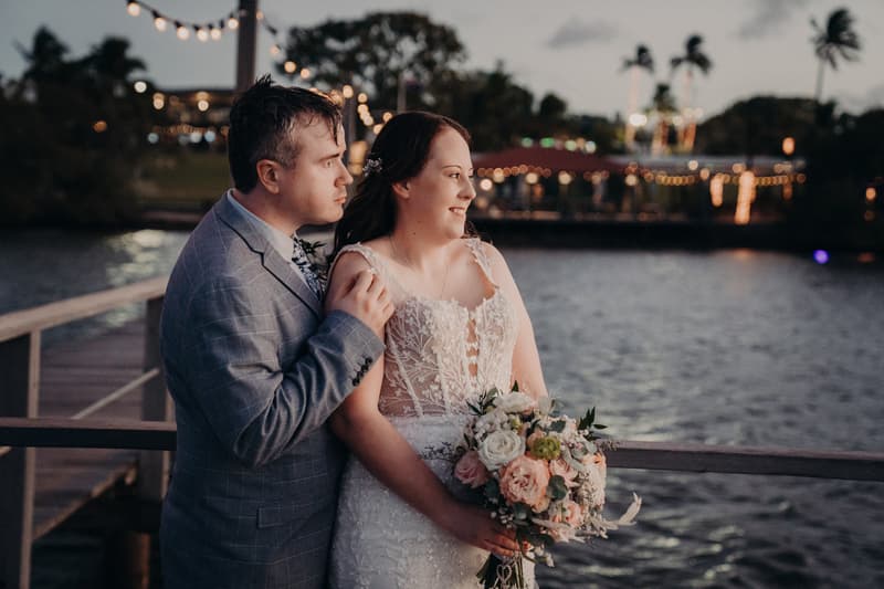Rebecca the bride and Dale the groom pose together on a wooden pier at Sandstone Point Hotel, with Rebecca holding a bouquet of flowers and twinkling lights visible in the background over the water.