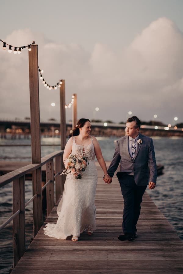 Rebecca the bride and Dale the groom walk hand-in-hand along a wooden pier at Sandstone Point Hotel, with Rebecca holding a bouquet and string lights overhead.