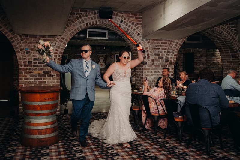 Rebecca and Dale enter the Sandstone Point Hotel — Cellar reception holding hands, with Rebecca raising a selfie stick and Dale holding a bouquet.