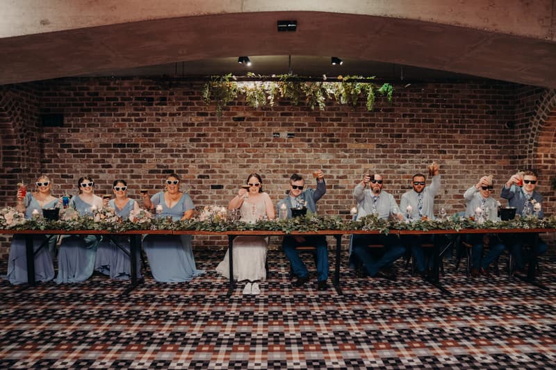 Rebecca the bride and Dale the groom sit at a long table with their bridal party at Sandstone Point Hotel — Cellar, all wearing sunglasses and raising drinks in a toast.