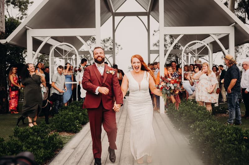 Bride Red and groom Jayden walk hand in hand down the aisle at Sandstone Point Hotel — The Pavilion, smiling and holding a bouquet, with guests standing and watching on both sides.