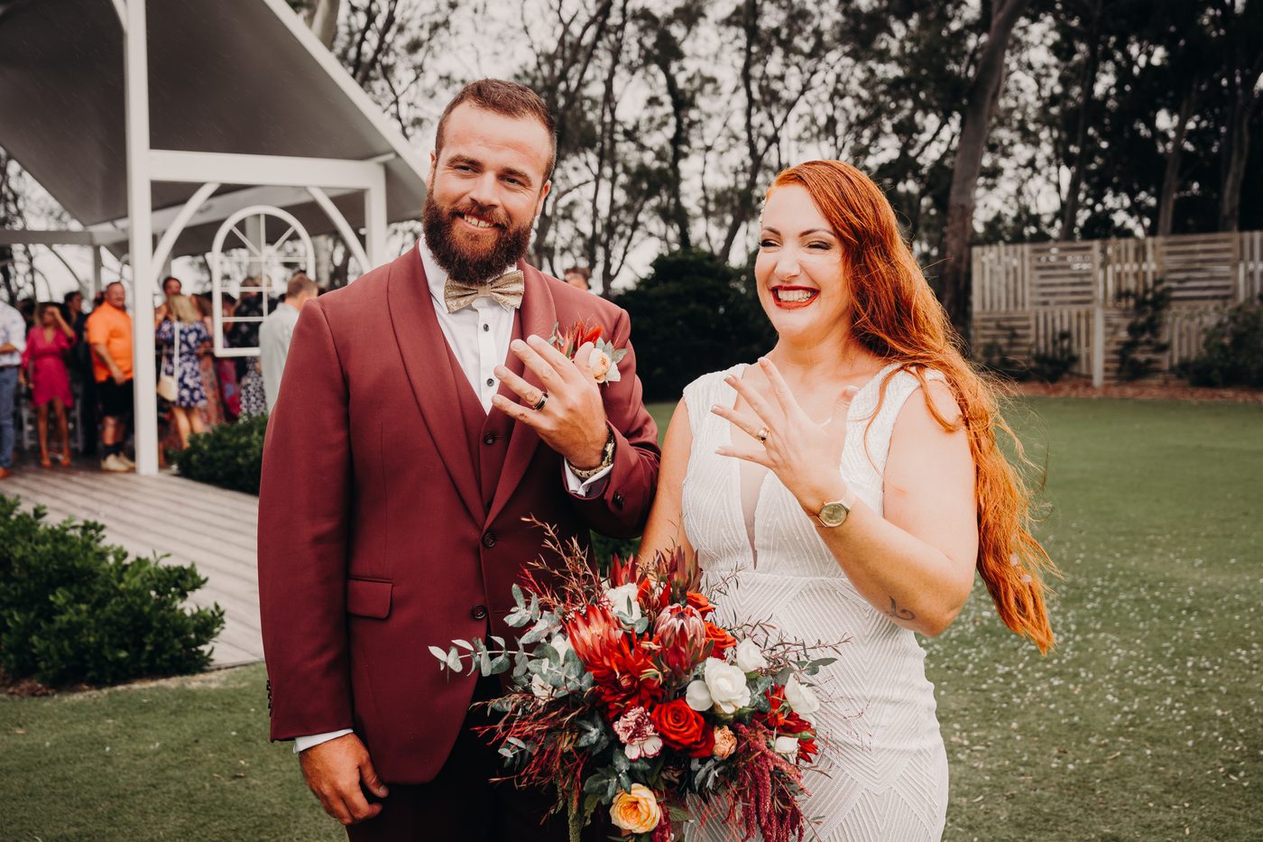 Bride Red and groom Jayden stand outside The Pavilion, with guests and a white gazebo structure visible in the background. Brisbane wedding photography by She Said Yes.