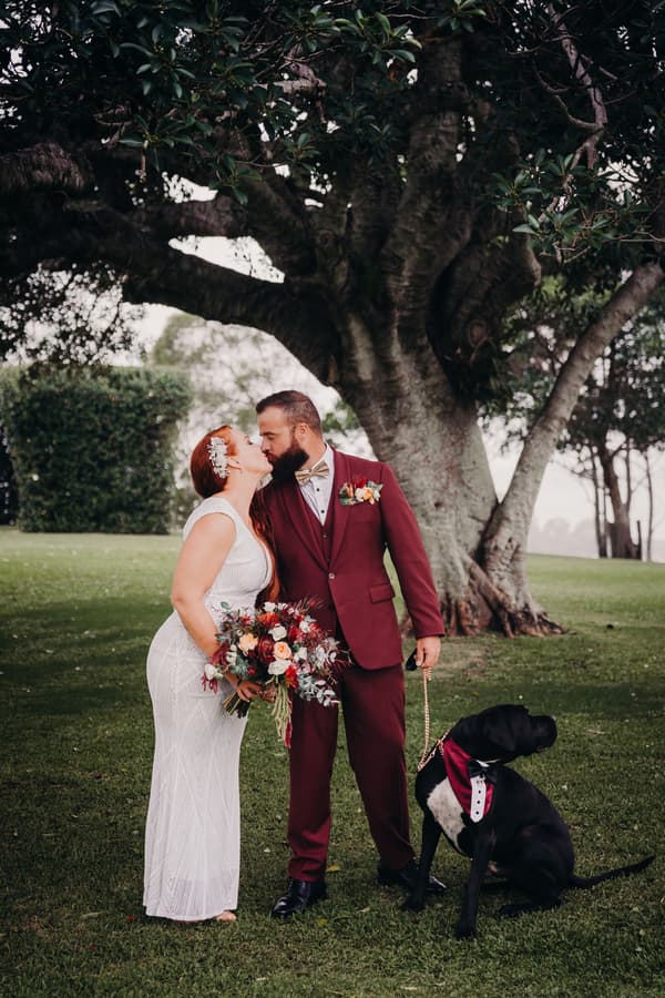 The bride and groom share a kiss outdoors under a large tree at Sandstone Point Hotel, The Pavilion. The bride holds a bouquet of flowers, and the groom holds a leash attached to a black dog wearing a red and white bow tie.
