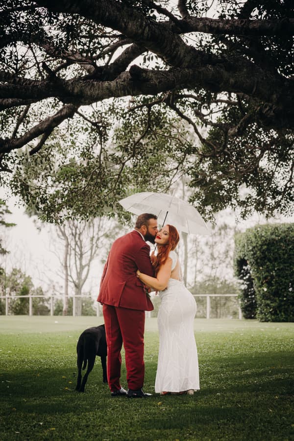 The bride in a white dress and the groom in a red suit embrace under a large tree while holding a transparent umbrella on a grassy area at Sandstone Point Hotel.