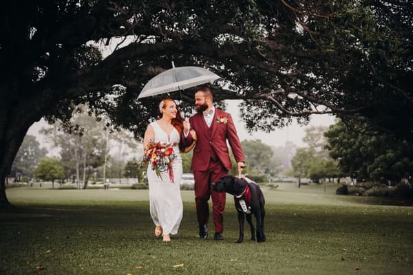 The bride Red and groom Jayden walk arm in arm under a large tree on a grassy lawn, with Jayden holding a transparent umbrella and a black dog wearing a tuxedo collar walking beside them. Elopement photography packages Brisbane by She Said Yes.