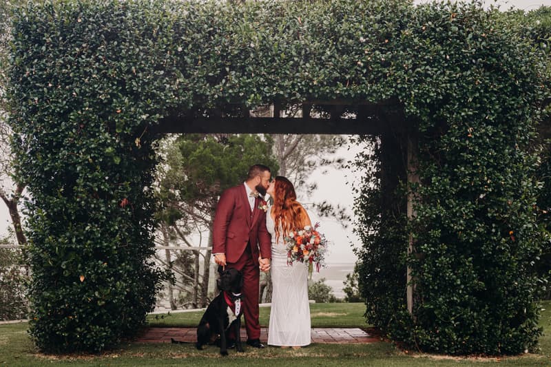 The bride and groom share a kiss while holding hands under a leafy arch at Sandstone Point Hotel, with their black dog sitting in front of them wearing a tuxedo collar.