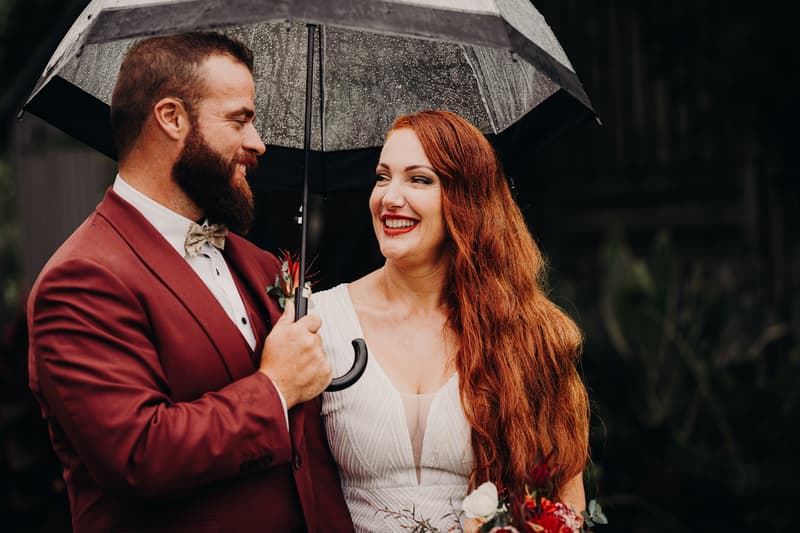 Red and Jayden stand close under a black umbrella with raindrops, Red holding a bouquet and smiling at Jayden.