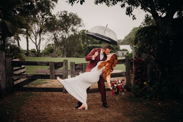 Jayden dips Red for a kiss outdoors near a wooden gate, holding a clear umbrella while Red holds a bouquet of flowers.