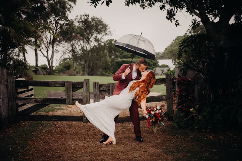 Jayden dips Red for a kiss outdoors near a wooden gate, holding a clear umbrella while Red holds a bouquet of flowers.