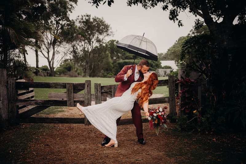 Jayden dips Red for a kiss outdoors near a wooden gate, holding a clear umbrella while Red holds a bouquet of flowers. Best wedding photographer Moreton Bay by She Said Yes.