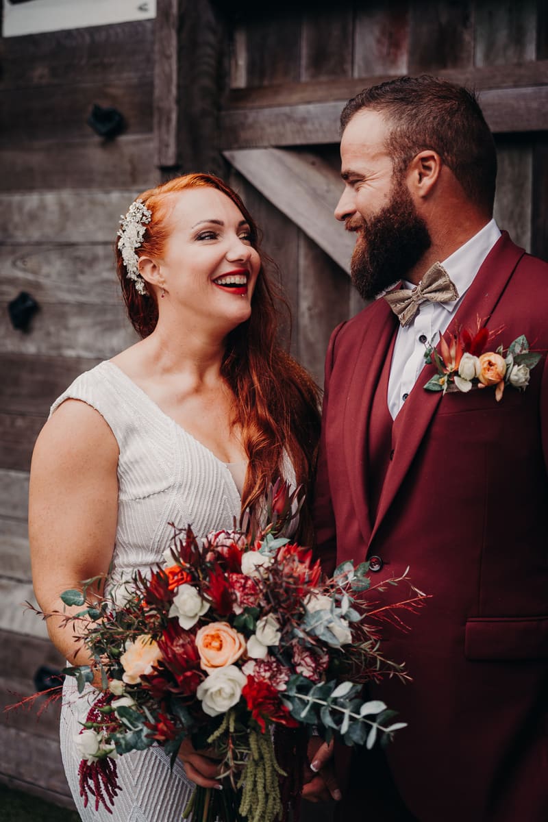 The bride Red and groom Jayden stand together in front of a wooden door, The Pavilion. Red holds a large bouquet of mixed flowers including red, white, and peach blooms. Jayden wears a burgundy suit with a bow tie and boutonniere. Ekka week Brisbane wedding by She Said Yes.