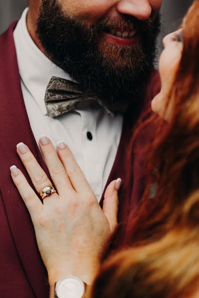 Close-up of the groom Jayden wearing a burgundy suit and patterned bow tie smiling as the bride Red places her hand with a ring on his chest. Elopement photographer Brisbane by She Said Yes.