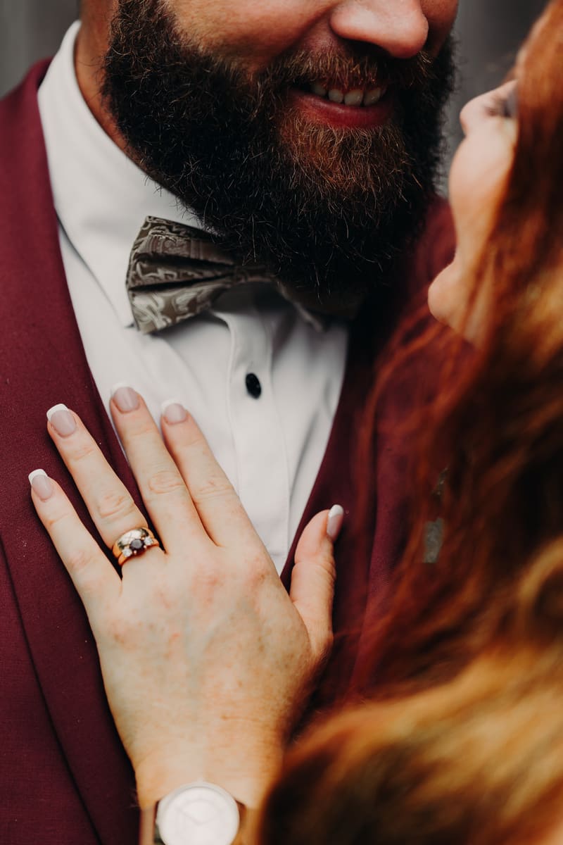 Close-up of the groom Jayden wearing a burgundy suit and patterned bow tie smiling as the bride Red places her hand with a ring on his chest. Wedding photography contracts by She Said Yes.