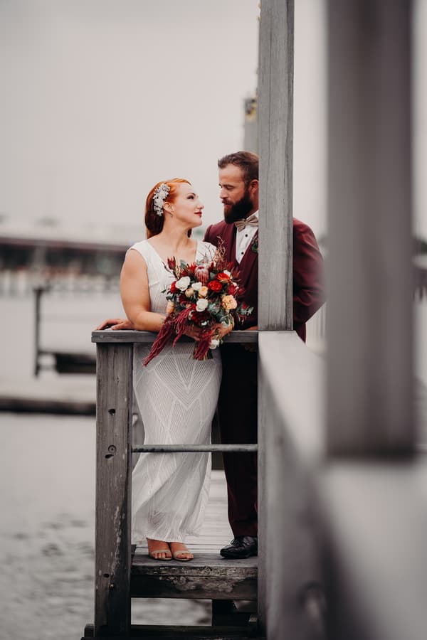 Red and Jayden stand closely together on a wooden dock by the water, with Red holding a bouquet of flowers and both looking at each other.