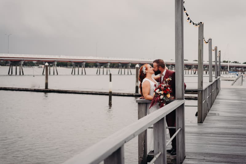 The bride and groom kiss on a wooden pier with string lights overhead and water in the background.