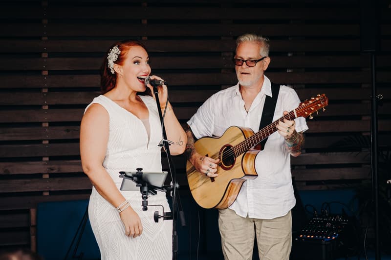 The bride sings into a microphone while a man plays acoustic guitar beside her at Sandstone Point Hotel — The Cellar.