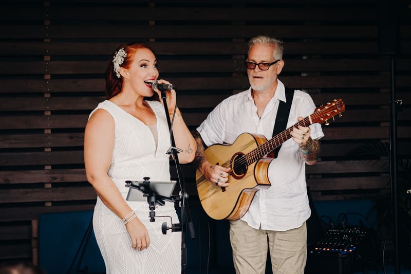 The bride sings into a microphone while a man plays acoustic guitar beside her. Cinematic videography wedding by She Said Yes.