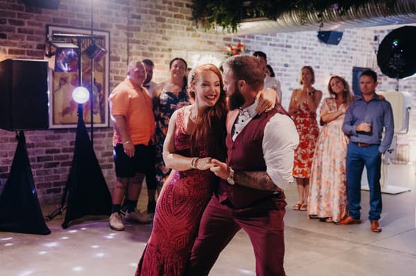The bride in a red dress and the groom in a burgundy vest and pants dance together at the reception stage of Sandstone Point Hotel — The Cellar, with guests watching in the background.
