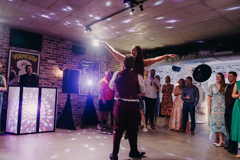 The groom Jayden lifts the bride Red with her arms outstretched on the dance floor at Sandstone Point Hotel — The Cellar, while guests watch and smile in the background near a DJ booth with colorful lights.