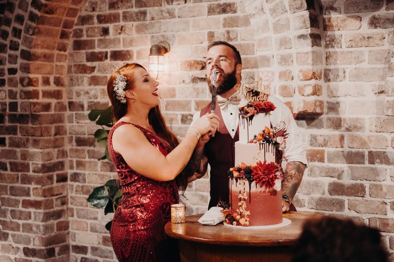 The bride in a red dress feeds a bite of wedding cake to the groom wearing a vest and bow tie at Sandstone Point Hotel — The Cellar reception stage.