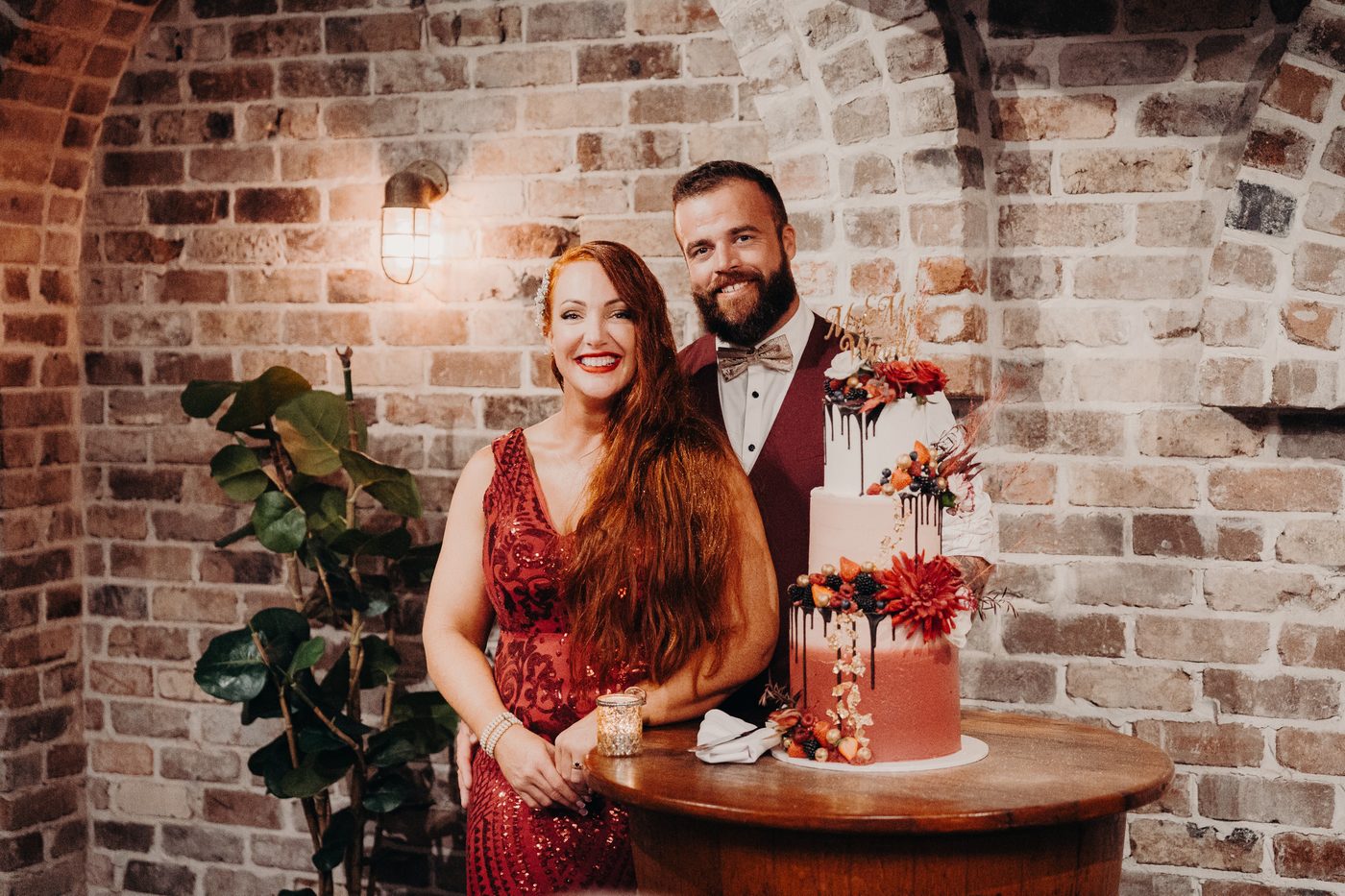 The bride in a red dress and the groom in a burgundy vest and bow tie stand behind a three-tier wedding cake decorated with flowers and berries. Wedding photography add-ons by She Said Yes.